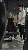 Young, attractive red-haired woman is exercising at gym using leg machine, lifting her legs with weights. Her fitness coach is standing next to her, monitoring her technique to ensure she is doing exercise correctly. video