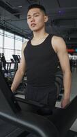 Attractive Asian man with a toned and muscular physique in a tank top is warming up on a treadmill before starting a workout at the gym. video