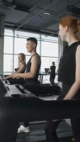 Three young friends are working out on treadmills at the gym, talking to each other and smiling while they do cardio. Vertical. video