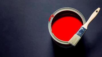 Open can of vibrant red paint with brush resting on edge against dark background, creating bold contrast. Perfect for painting services, interior design, color trends, DIY and creative branding photo