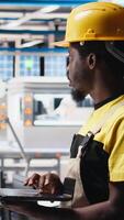 Vertical Black technician in solar panel facility reviews photovoltaic system performance on laptop. Specialist analyzing production line metrics to optimize efficiency in industrial plant. Camera B. video