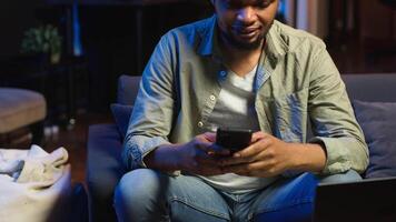Carefree freelancer chatting with friends over text messages on phone, sitting on the couch and scrolling social media apps. Black guy showcases staying entertained while working remotely. Camera B. video