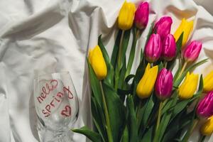 Hello SPRING message on wine glass with Multicolored fresh tulips on white bed with trendy deep shadows. Spring flowers in yellow and violet colors. Romantic postcard wallpaper design Copy space photo