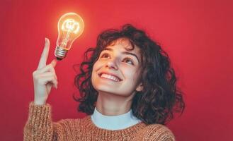 Studio portrait of a young woman holding a glowing light bulb on a red background, representing a new idea or solution photo