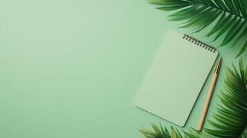 Green notebook and pen on a light green surface. Palm leaves decorate the scene. Overhead shot with empty space to the left. photo