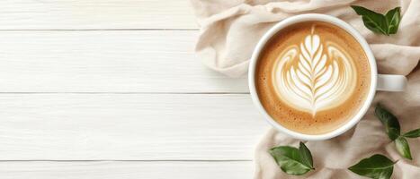 Coffee cup with latte art on white wood table. Beige cloth and green leaves surround the coffee. Overhead view of beverage on wooden surface photo