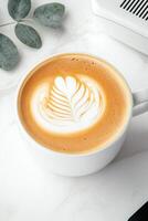 Close up of latte art coffee in white cup. Leaf design in foam. On white marble table with green leaves. Top view photo