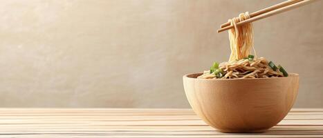 Noodles in wood bowl. Chopsticks lifting noodles. Food is on wood table. Simple beige background. Delicious looking meal with green vegetable photo
