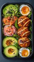 Healthy lunch box with avocado, eggs, salmon, and leafy greens on a dark background. Colorful and nutritious meal in a black container photo