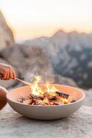 Burning firewood in a stone fire pit. Flames and smoke rising against a backdrop of blurred mountains during sunset. Hand holding a tool photo