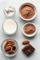 Dessert ingredients arrangement. Chocolate brownie square with pecans. Bowls hold cocoa, milk, flour, chips. Gray background. Preparing vegetarian desserts. photo