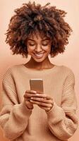 Smiling woman with curly hair types on mobile device. Happy business owner wears glasses and beige sweater over plain background. photo