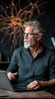 Man sits at table using laptop. Neural network style background. Gray hair, beard, glasses. Orange neuron connections on wall behind. photo