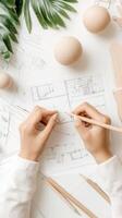 Hands hold pencil above sketch on paper. Green leaves and wooden objects decorate white background. Lines and geometric shapes describe architectural structure. photo