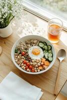 Healthy breakfast bowl with egg grains vegetables and chickpeas sits on a table near a window with natural light and a beverage photo