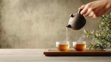 Hand pouring hot tea from brown teapot into clear glass cups on a wooden tray with plant against grey textured background photo
