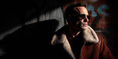 A man in a brown coat and sunglasses standing in front of a wall photo