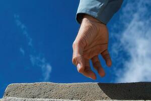 A hand reaching out towards a stone ledge against a bright blue sky with clouds photo