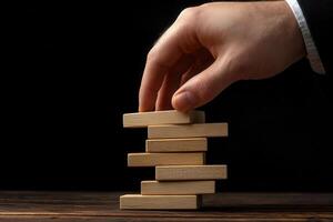 Hand carefully placing a wooden block on a tower of blocks against a dark background photo
