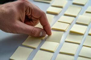 Hand selecting a sticky note from a grid of yellow notes on a white surface, focused work photo