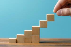 Hand placing wooden block on a growing staircase of blocks against a blue background photo