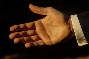 Open hand gesture in a formal setting, symbolizing invitation or agreement. Background shows blurred dark tones photo