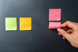 Hand placing a pink sticky note on a black surface alongside green, yellow notes photo