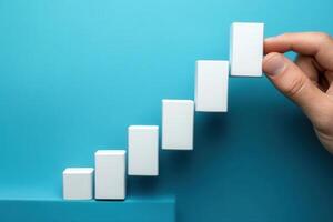Hand placing a white block on a staircase of blocks against a blue background, symbolizing growth photo