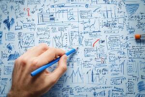 Hand holding blue pencil writing on chaotic paper filled with graphs and notes photo