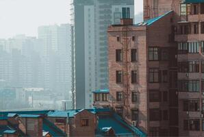 Brick buildings with blue roofs in the foreground contrast with a hazy cityscape of modern skyscrapers. Urban architecture blends old and new under a foggy sky. photo