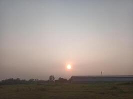 A field with a barn and a sun setting behind it photo