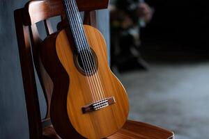 Classical guitar resting on wooden chair. Wooden guitar body, strings, and fretboard are visible. Dark background photo