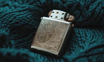 Silver lighter with intricate gold-toned design, resting on a teal knitted fabric photo