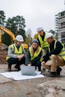 Construction team reviewing blueprints on-site. Several construction workers in safety vests and hard hats, huddled around a laptop and blueprints, focused on project plans on a construction site photo