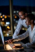 Two colleagues collaborate late at night on a computer, reviewing data or a presentation. City lights visible outside the window photo