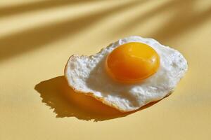 Single fried egg on a pale yellow surface, sunlight casts shadows. The egg is cooked, with a runny yolk photo