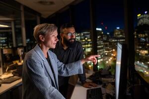 Two colleagues collaborate late at night on a project, reviewing a computer screen. City lights visible through large office windows photo