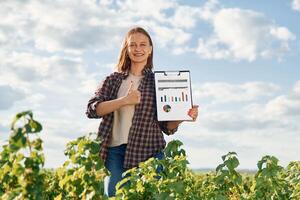 Document with graphs and plan in hands. Young woman is on the beautiful agricultural field at daytime photo