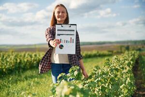 Document with graphs and plan, in hands. Young woman is on the beautiful agricultural field at daytime photo
