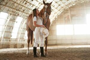 Daylight comes through the ,windows. Beautiful young woman is with horse indoors photo