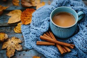 Blue mug with hot drink, cinnamon sticks and knitted sweater on rustic table with autumn leaves in warm colors photo