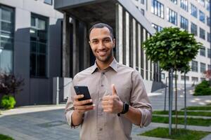 Optimistic muslim male showing thumb up gesture while using smartphone and smiling at camera outdoors. Proud guy receiving pleasant notification about stock prices rising while standing at downtown. photo