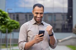 Young muslim male businessman standing on street near office building and using mobile phone, looking at screen and happy with received news and message showing victory gesture with hand. photo