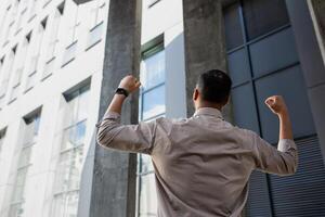 Back view of brunet man in shirt making power sign with clenched fists while standing next to office building. Unrecognizable guy feeling strong and powerful after getting hired for dream job. photo
