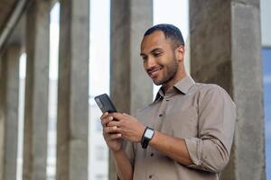 Carefree middle eastern man with digital watch on hand looking at smartphone screen on concrete columns background. Unshaved man using internet for searching new spots and routes for walk outdoors. photo