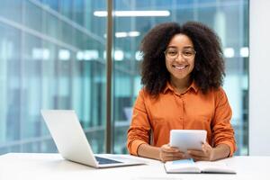 A smiling woman with glasses holds a tablet while working in a modern office environment. A laptop and notebook are on the desk. photo