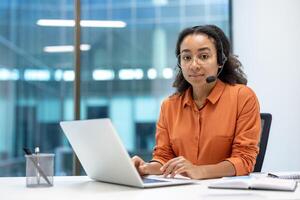 Portrait of serious focused woman with headset, customer support worker inside office at workplace, using laptop for call. Looking thoughtfully at camera. photo