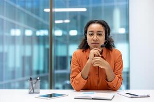 A young woman with a headset sits at a desk in an office setting, looking directly at the camera. She is ready to assist clients. photo