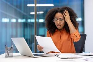 A businesswoman expresses worry while looking at a document. She is seated at a desk with a laptop, papers, and a pen holder in front of her. photo