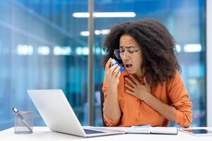 An office worker is experiencing an asthma attack. She struggles to breathe while reaching for her inhaler near her open laptop and notebook. photo